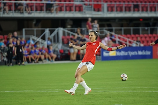 A soccer player in a red jersey prepares to kick a soccer ball on a green field. The background features spectators sitting in red seats, and a sign displaying 'Sports Medicine' is visible.