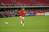 A female soccer player in a red jersey and white shorts is running towards a soccer ball on a grassy field. The stadium seats are mostly empty, with a few spectators visible in the background. Advertising banners are displayed along the stands.