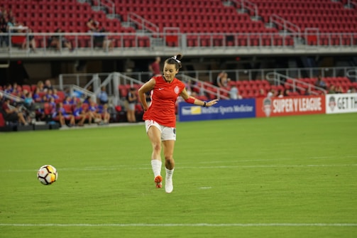 A female soccer player in a red jersey and white shorts is running towards a soccer ball on a grassy field. The stadium seats are mostly empty, with a few spectators visible in the background. Advertising banners are displayed along the stands.