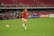 A female soccer player in a red jersey and white shorts is running towards a soccer ball on a grassy field. The stadium seats are mostly empty, with a few spectators visible in the background. Advertising banners are displayed along the stands.