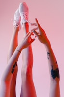 Two feet wearing white mesh sneakers are raised against a pink background. Hands with tattoos and painted nails are reaching up towards the feet.