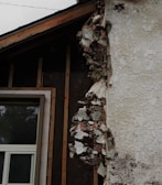A section of a building with exposed and damaged wall layers is visible. The surface shows crumbling plaster revealing the brick and insulation beneath. A window with a white frame is partially seen, surrounded by unfinished wooden beams.