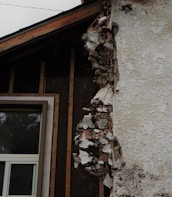 A section of a building with exposed and damaged wall layers is visible. The surface shows crumbling plaster revealing the brick and insulation beneath. A window with a white frame is partially seen, surrounded by unfinished wooden beams.