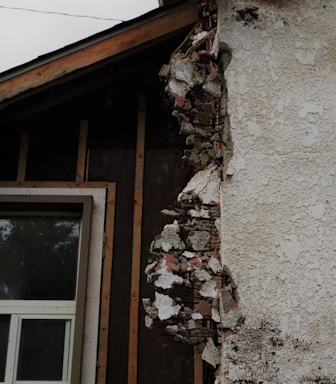 A section of a building with exposed and damaged wall layers is visible. The surface shows crumbling plaster revealing the brick and insulation beneath. A window with a white frame is partially seen, surrounded by unfinished wooden beams.