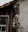 A section of a building with exposed and damaged wall layers is visible. The surface shows crumbling plaster revealing the brick and insulation beneath. A window with a white frame is partially seen, surrounded by unfinished wooden beams.