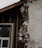 A section of a building with exposed and damaged wall layers is visible. The surface shows crumbling plaster revealing the brick and insulation beneath. A window with a white frame is partially seen, surrounded by unfinished wooden beams.
