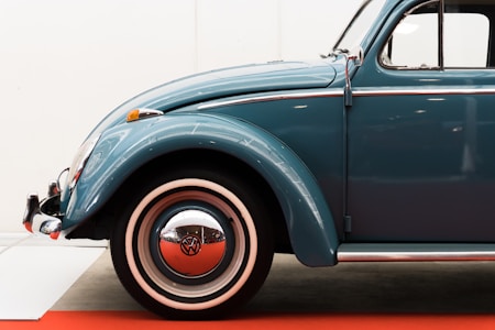 A classic vintage car with a round headlight, chrome accents, and a blue body is parked against a white backdrop, standing out against a red carpeted floor. The iconic design includes whitewall tires and distinctive Volkswagen branding on the wheel hub.