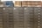 A row of gray metal filing cabinets with drawers, each labeled with small white and black tags. On top of the cabinets, there is a collection of hardbound books arranged in two groups. The books are a mix of blue and brown colors, and they appear to be reference or index volumes.