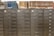 A row of gray metal filing cabinets with drawers, each labeled with small white and black tags. On top of the cabinets, there is a collection of hardbound books arranged in two groups. The books are a mix of blue and brown colors, and they appear to be reference or index volumes.