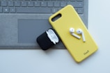 A yellow phone case with wireless earbuds sits on top of a laptop keyboard. An open black charging case for the earbuds is nearby.