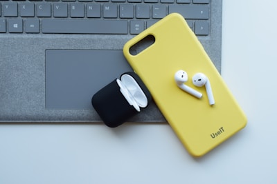 A yellow phone case with wireless earbuds sits on top of a laptop keyboard. An open black charging case for the earbuds is nearby.