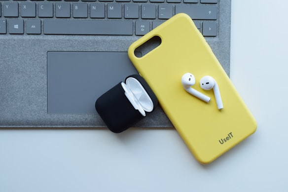 A yellow phone case with wireless earbuds sits on top of a laptop keyboard. An open black charging case for the earbuds is nearby.