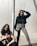 Two women are posed against a modern metallic background. One woman is squatting, wearing a beige jacket and black pants, while the other stands confidently with her hand in her hair, wearing a teal fur jacket and gray pants. The sunlight casts distinct shadows across the scene.