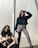 Two women are posed against a modern metallic background. One woman is squatting, wearing a beige jacket and black pants, while the other stands confidently with her hand in her hair, wearing a teal fur jacket and gray pants. The sunlight casts distinct shadows across the scene.
