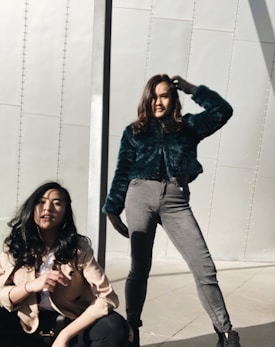 Two women are posed against a modern metallic background. One woman is squatting, wearing a beige jacket and black pants, while the other stands confidently with her hand in her hair, wearing a teal fur jacket and gray pants. The sunlight casts distinct shadows across the scene.