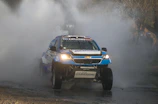 The rally car crossing a shallow river with water splashing dramatically around the wheels.