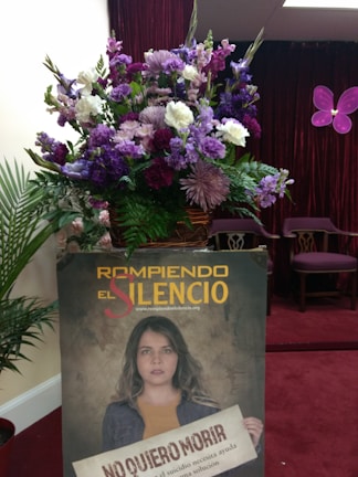 A large bouquet of flowers with purple, white, and pink blossoms is placed in a pot against a backdrop of a deep red curtain. Next to it is a poster featuring a person holding a sign with text about suicide prevention. There is a stylized purple butterfly decoration on the wall.