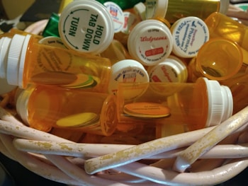A collection of empty orange prescription pill bottles with white childproof caps is inside a worn wicker basket. Each cap displays various pharmacy labels and instructions. The surrounding setting is dimly lit, highlighting the details on the bottles and basket.