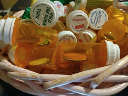 A collection of empty orange prescription pill bottles with white childproof caps is inside a worn wicker basket. Each cap displays various pharmacy labels and instructions. The surrounding setting is dimly lit, highlighting the details on the bottles and basket.