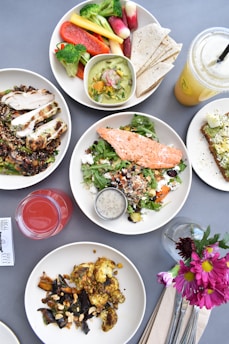 A variety of healthy dishes presented on a table. There is a plate with a grilled salmon salad, another with grilled chicken and quinoa, and another plate with roasted vegetables. A bowl of assorted fresh vegetables and a side of tortillas can be seen, along with two beverages, one red and one yellow. A piece of avocado toast garnished with toppings is also present. Flowers in a small vase add a decorative touch.