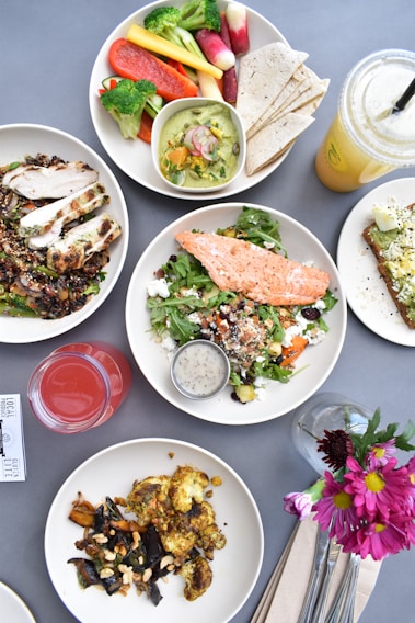 A variety of healthy dishes presented on a table. There is a plate with a grilled salmon salad, another with grilled chicken and quinoa, and another plate with roasted vegetables. A bowl of assorted fresh vegetables and a side of tortillas can be seen, along with two beverages, one red and one yellow. A piece of avocado toast garnished with toppings is also present. Flowers in a small vase add a decorative touch.