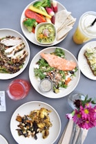 A variety of healthy dishes presented on a table. There is a plate with a grilled salmon salad, another with grilled chicken and quinoa, and another plate with roasted vegetables. A bowl of assorted fresh vegetables and a side of tortillas can be seen, along with two beverages, one red and one yellow. A piece of avocado toast garnished with toppings is also present. Flowers in a small vase add a decorative touch.