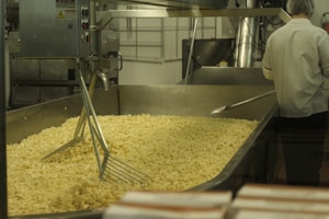A large industrial food processing setup with a metal container holding a significant amount of pasta. A worker in a white uniform and hairnet is seen from the back, attending to the machinery or pasta. The environment appears to be a clean industrial kitchen.