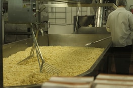 A large industrial food processing setup with a metal container holding a significant amount of pasta. A worker in a white uniform and hairnet is seen from the back, attending to the machinery or pasta. The environment appears to be a clean industrial kitchen.