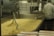 A large industrial food processing setup with a metal container holding a significant amount of pasta. A worker in a white uniform and hairnet is seen from the back, attending to the machinery or pasta. The environment appears to be a clean industrial kitchen.