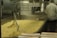 A large industrial food processing setup with a metal container holding a significant amount of pasta. A worker in a white uniform and hairnet is seen from the back, attending to the machinery or pasta. The environment appears to be a clean industrial kitchen.