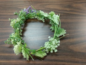 A handmade floral wreath lies on a wooden surface. It consists of various green stems intertwined with small white and light purple flowers. The arrangement is circular and appears freshly picked.