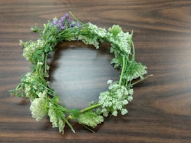 A handmade floral wreath lies on a wooden surface. It consists of various green stems intertwined with small white and light purple flowers. The arrangement is circular and appears freshly picked.