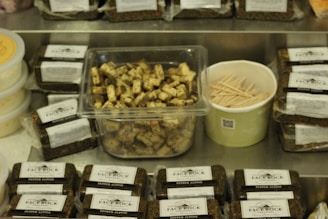 A variety of packaged cheese blocks labeled 'Face Rock Pepper Alpine' are displayed on a metal surface. In the center, a clear container holds cubed cheese pieces with visible herbs. Beside it, a green container filled with wooden toothpicks. Containers of a spreadable substance are on the left.