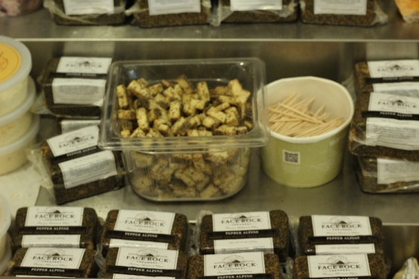 A variety of packaged cheese blocks labeled 'Face Rock Pepper Alpine' are displayed on a metal surface. In the center, a clear container holds cubed cheese pieces with visible herbs. Beside it, a green container filled with wooden toothpicks. Containers of a spreadable substance are on the left.