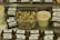 A variety of packaged cheese blocks labeled 'Face Rock Pepper Alpine' are displayed on a metal surface. In the center, a clear container holds cubed cheese pieces with visible herbs. Beside it, a green container filled with wooden toothpicks. Containers of a spreadable substance are on the left.