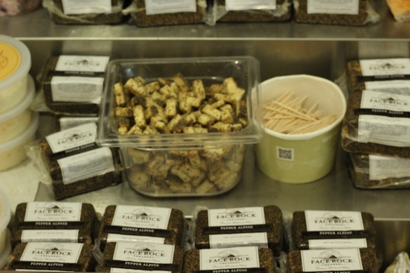 A variety of packaged cheese blocks labeled 'Face Rock Pepper Alpine' are displayed on a metal surface. In the center, a clear container holds cubed cheese pieces with visible herbs. Beside it, a green container filled with wooden toothpicks. Containers of a spreadable substance are on the left.