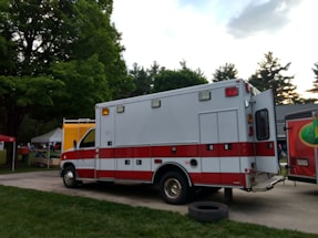 A red and white ambulance is parked on a paved area near a grassy field. The vehicle is positioned with its rear end facing towards the viewer and the back doors are open. Surrounding the ambulance are trees and tents, likely indicating an outdoor event or festival setting. A tire lies on the ground nearby.