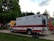 A red and white ambulance is parked on a paved area near a grassy field. The vehicle is positioned with its rear end facing towards the viewer and the back doors are open. Surrounding the ambulance are trees and tents, likely indicating an outdoor event or festival setting. A tire lies on the ground nearby.