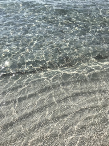 Clear, shallow water with visible ripples and patterns created by sunlight reflecting on the sandy seabed. The sunlight creates sparkling effects on the water surface, giving a sense of tranquility and natural beauty.