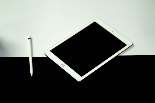 A tablet with a blank black screen lies on a contrasting surface divided between white and black. To the left of the tablet is a white stylus, placed on the white section of the surface.