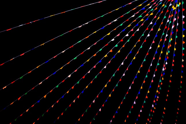 Colorful triangular flags are strung across a dark background in parallel lines, creating a vibrant and festive atmosphere. The flags are arranged in various colors such as red, blue, green, yellow, and pink.