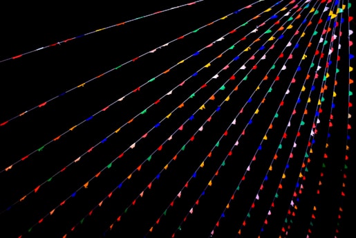 Colorful triangular flags are strung across a dark background in parallel lines, creating a vibrant and festive atmosphere. The flags are arranged in various colors such as red, blue, green, yellow, and pink.