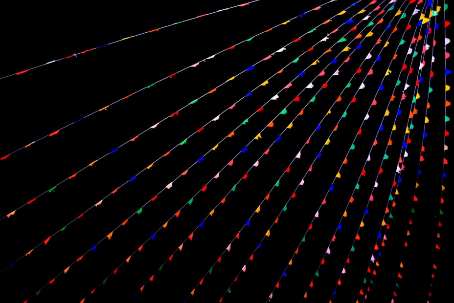 Colorful triangular flags are strung across a dark background in parallel lines, creating a vibrant and festive atmosphere. The flags are arranged in various colors such as red, blue, green, yellow, and pink.