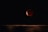 A dark night sky with a prominent red moon or lunar eclipse. The moon appears partially shadowed, casting a reddish hue. There are faint stars visible in the background. The bottom part of the image shows a wooden surface or horizon line.