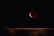 A dark night sky with a prominent red moon or lunar eclipse. The moon appears partially shadowed, casting a reddish hue. There are faint stars visible in the background. The bottom part of the image shows a wooden surface or horizon line.