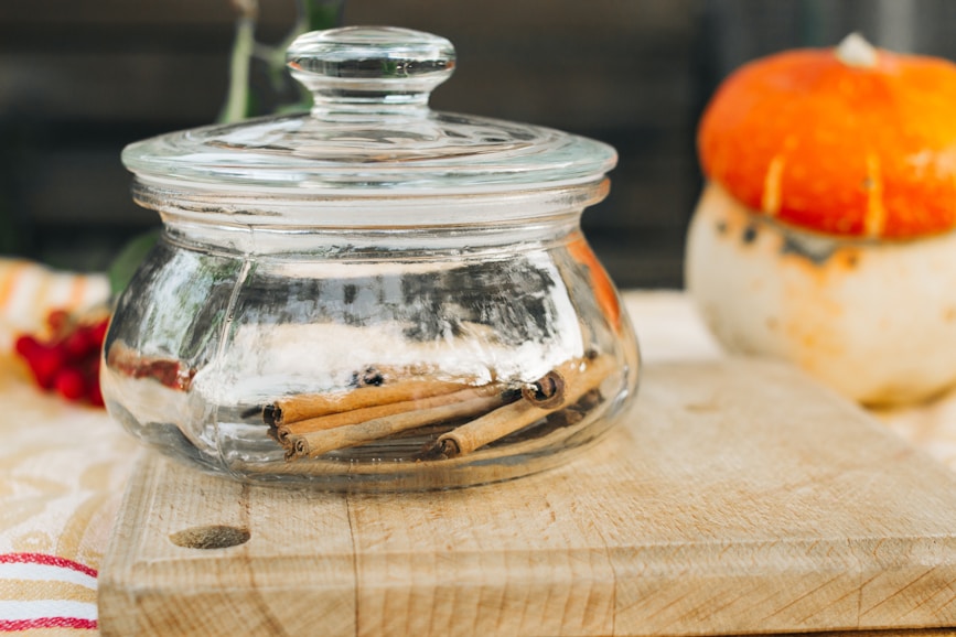 A glass jar with a lid contains cinnamon sticks, sitting on a wooden cutting board. A decorative orange gourd with cloves pressed into its surface is nearby. The background includes blurred red berries and striped cloth, suggesting an autumnal or rustic theme.