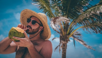 A person is enjoying a drink from a coconut using a straw while wearing sunglasses and a straw hat. They are standing under a palm tree with a bright blue sky in the background.