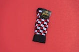 A pair of colorful patterned socks with a geometric cube design featuring red, black, and white colors is placed on a solid red background. The socks are neatly folded, and there is a cardboard label attached near the top bearing the brand name.