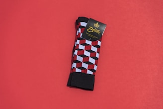 A pair of colorful patterned socks with a geometric cube design featuring red, black, and white colors is placed on a solid red background. The socks are neatly folded, and there is a cardboard label attached near the top bearing the brand name.