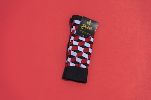 A pair of colorful patterned socks with a geometric cube design featuring red, black, and white colors is placed on a solid red background. The socks are neatly folded, and there is a cardboard label attached near the top bearing the brand name.